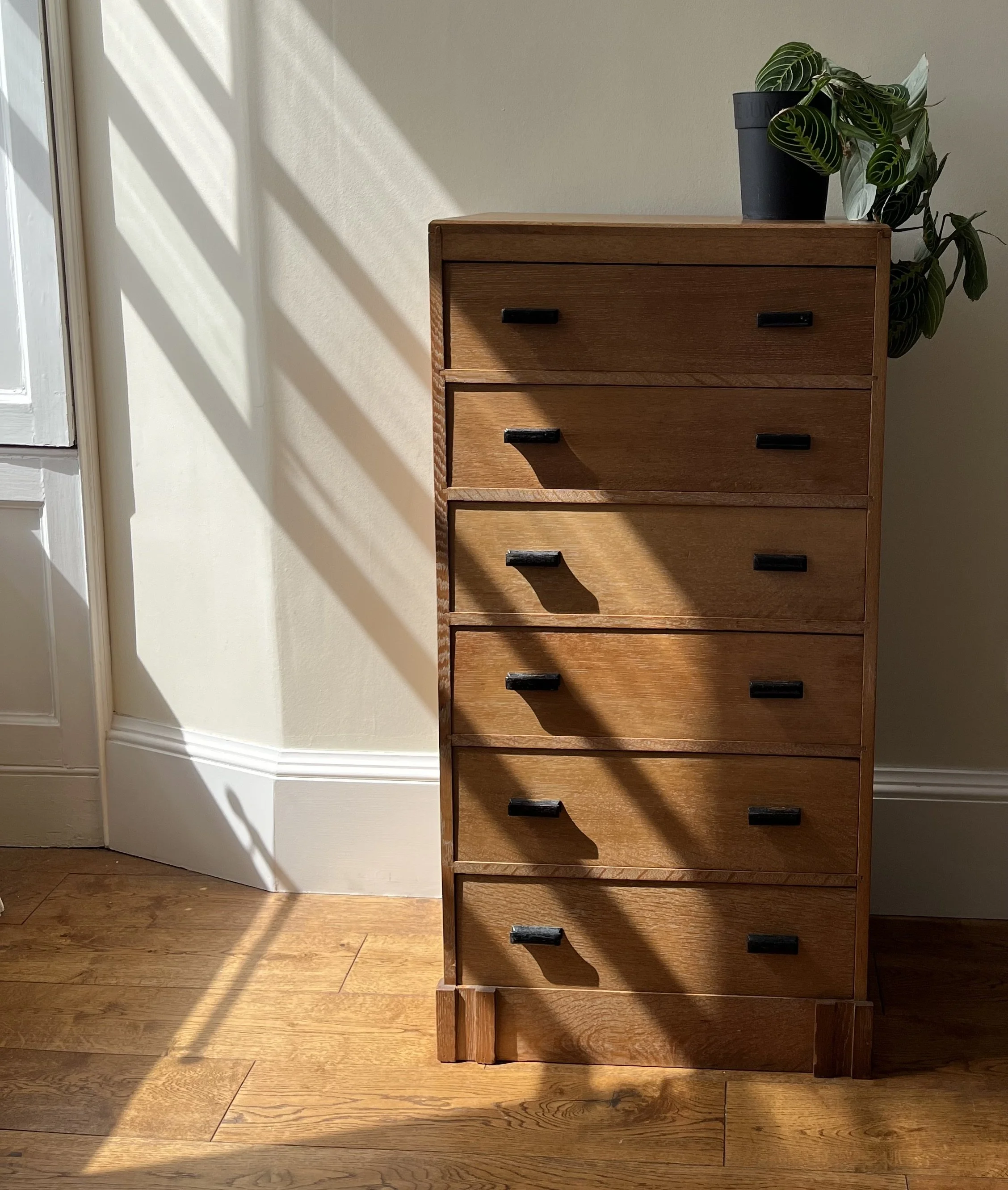 A wooden dresser with black handles standing against a white wall, with a potted plant on top, and sunlight casting shadows on the floor and wall.
