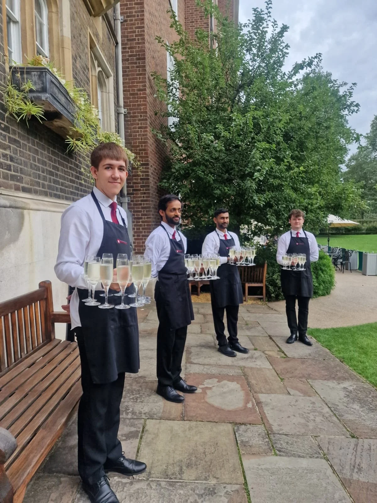 Four men dressed as waiters standing outdoors with trays of champagne glasses, a building with brick walls, a tree, and a grassy area in the background.
