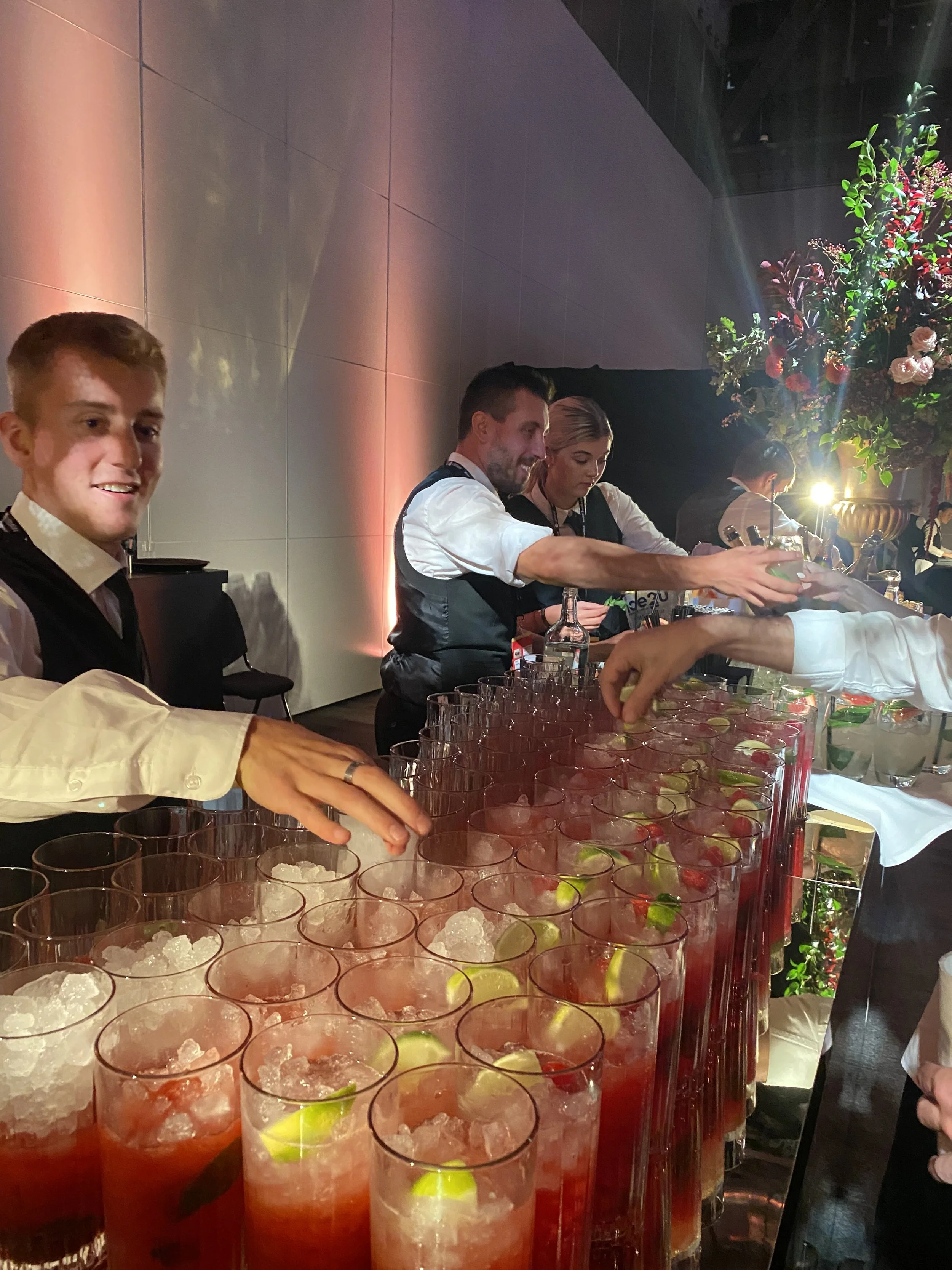 Bar with bartenders preparing colorful cocktails with lime wedges and ice at an event.