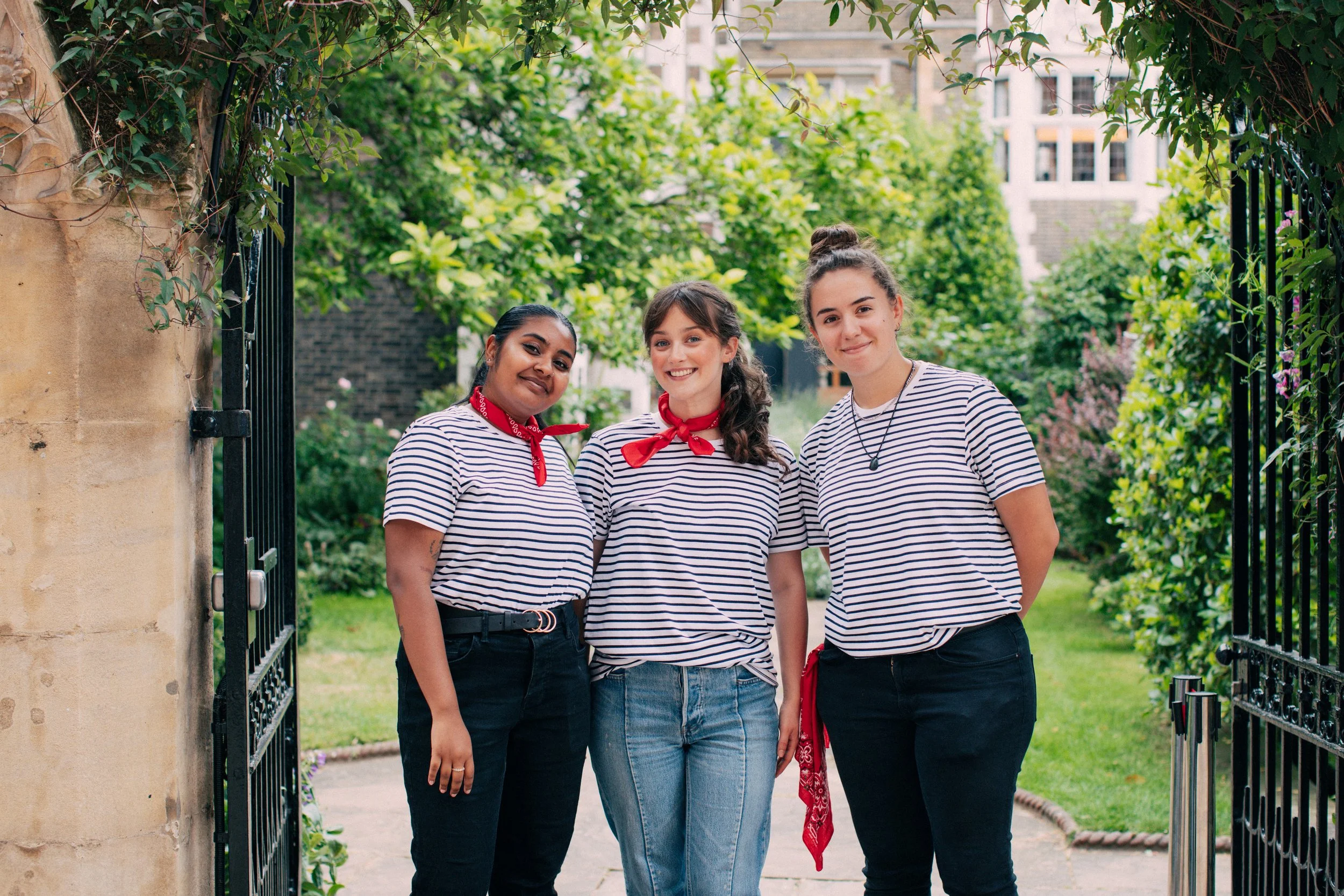 Three women standing together outdoors, wearing matching striped shirts and red neck scarves, smiling at the camera amidst green trees and plants.