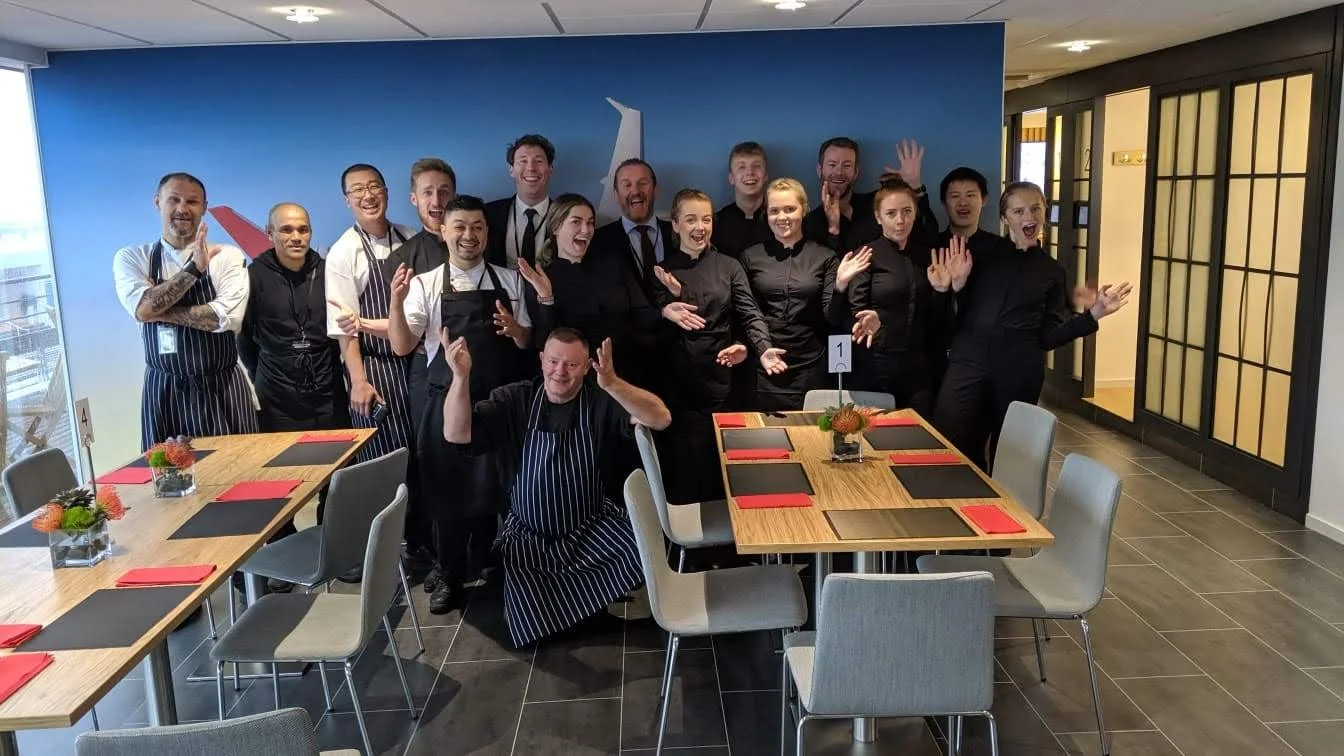 Group of restaurant staff members, including chefs and waitstaff, posing inside a restaurant, smiling and making gestures of celebration.
