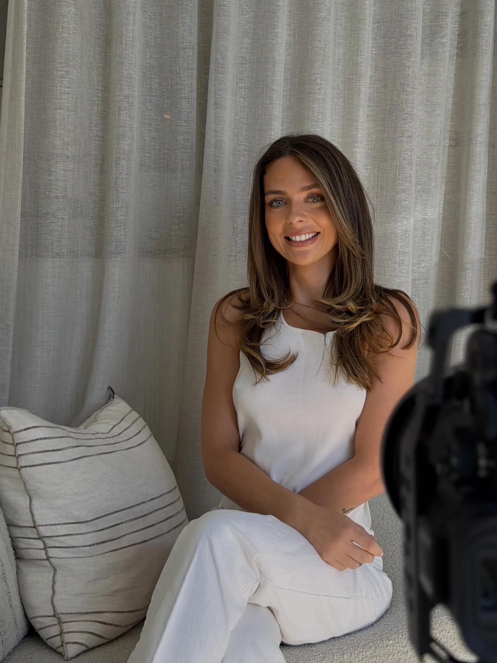 A woman with long brown hair wearing a sleeveless white top and white pants smiling at the camera while sitting on a light-colored sofa with a striped pillow behind her in front of a beige curtain.