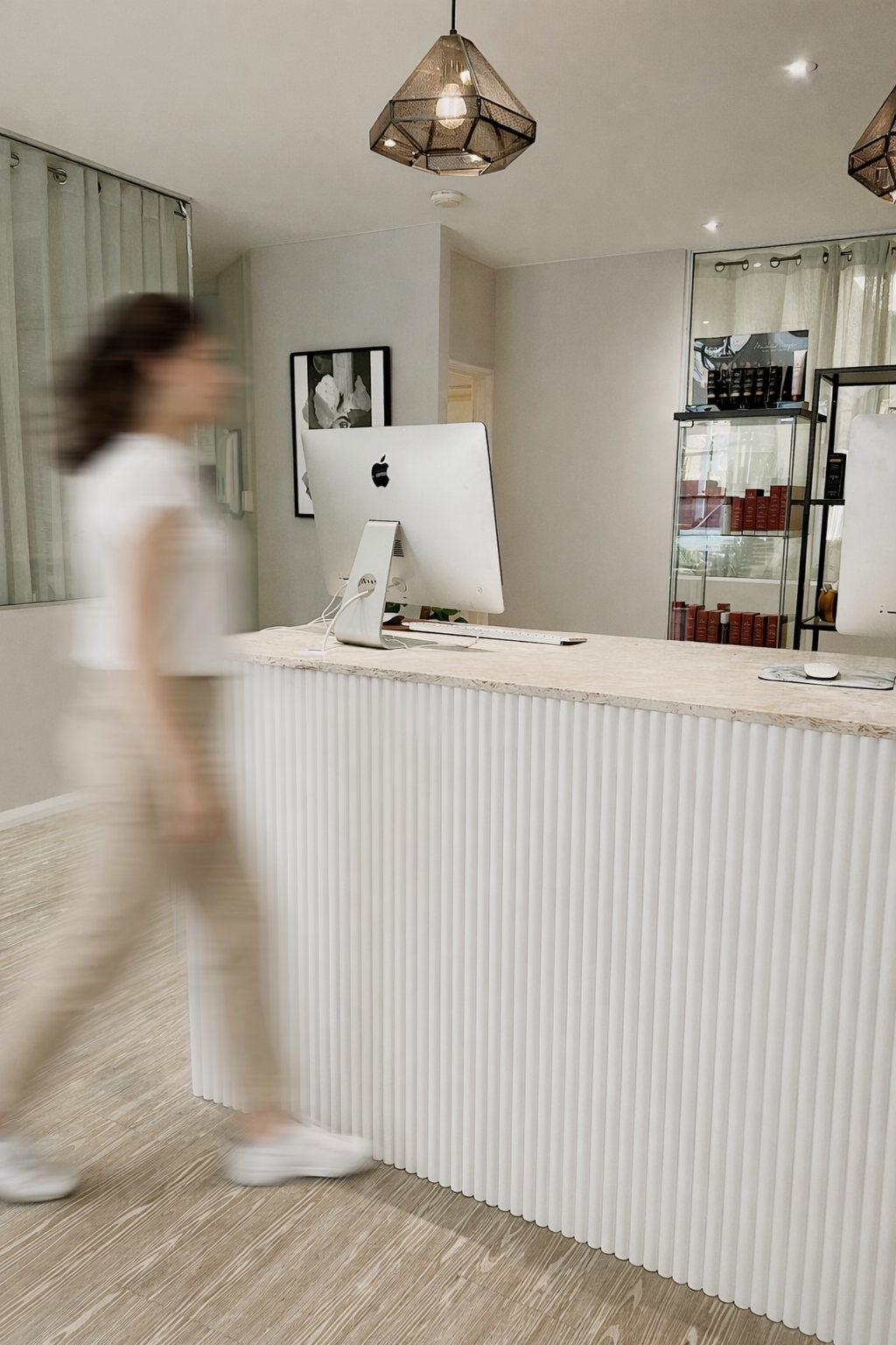 Blurry woman walking past a reception desk with an iMac computer in a modern office or reception area.