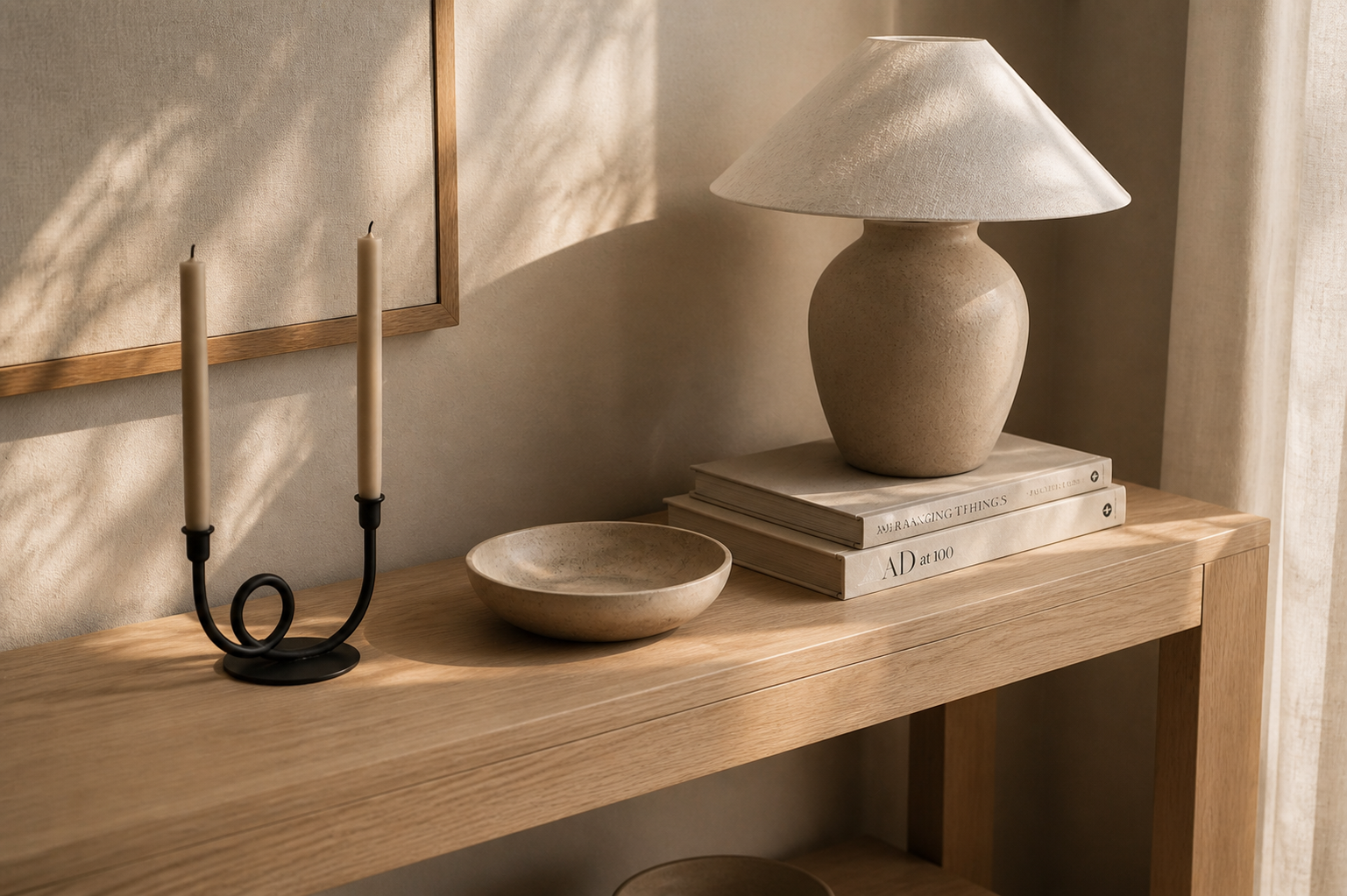 Mesa de madera con candelabro de dos velas, plato, pila, y libros con lámpara en fondo de pared beige.