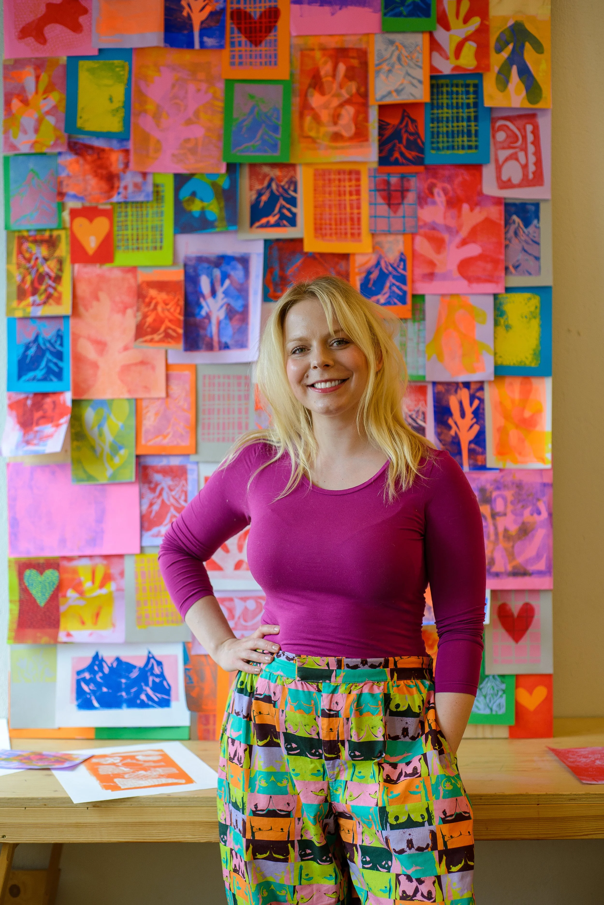 A woman with blonde hair wearing a magenta long-sleeve shirt and colorful pants stands in front of a vibrant wall of abstract artwork. She is smiling with her hand on her hip.