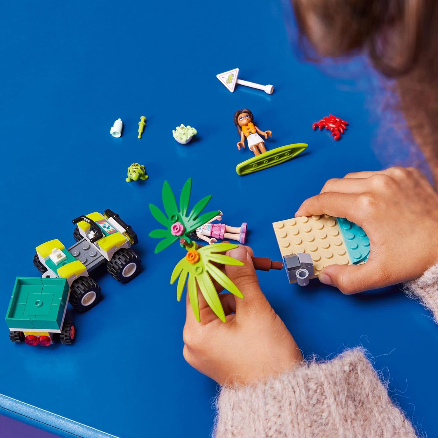 Child assembling a LEGO set on a blue table, with LEGO figures, vehicles, and accessories scattered around.