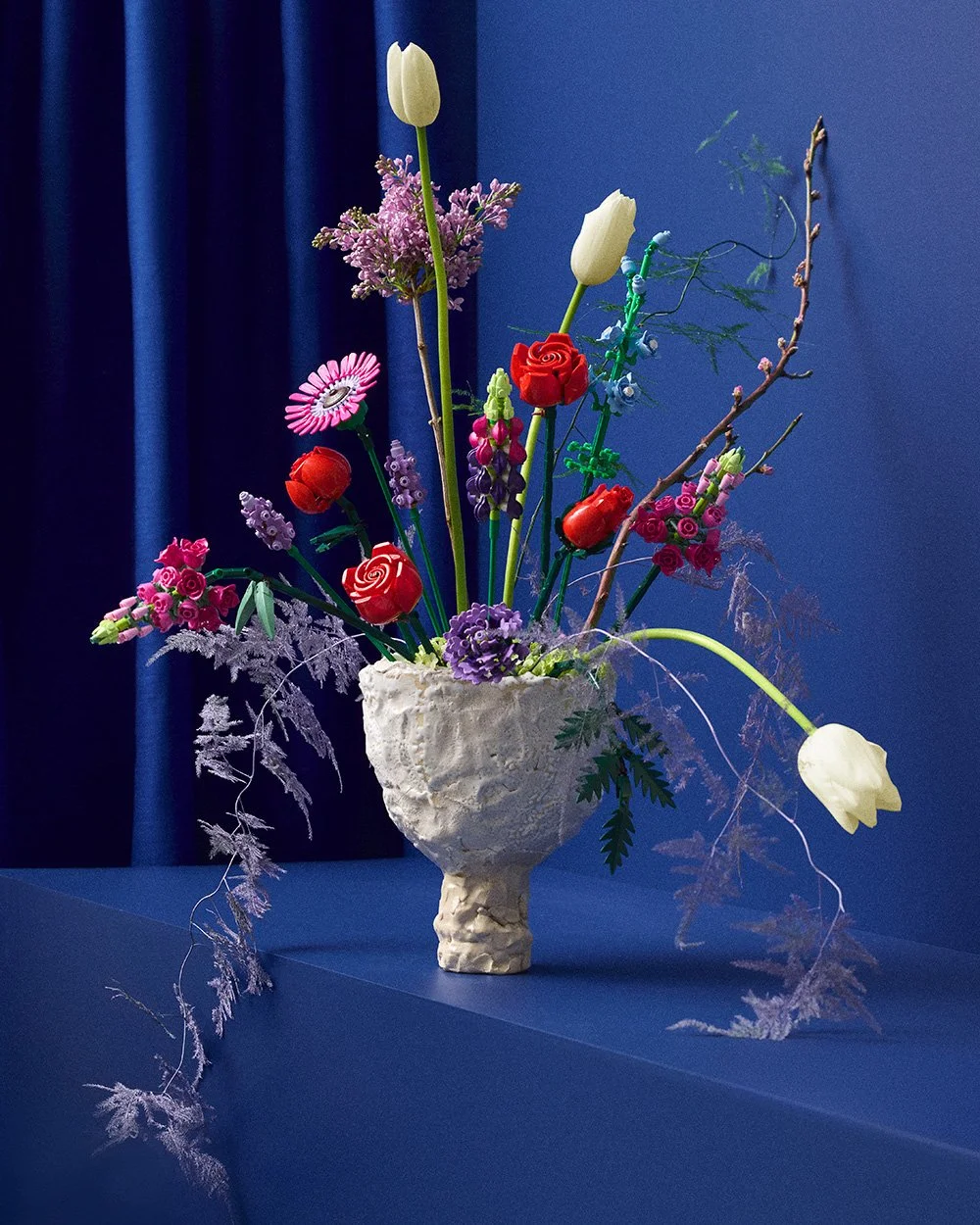 Arranged artificial flower bouquet in a textured cream-colored vase, featuring white tulips, red roses, purple flowers, and various green and pink accents on a dark blue background.