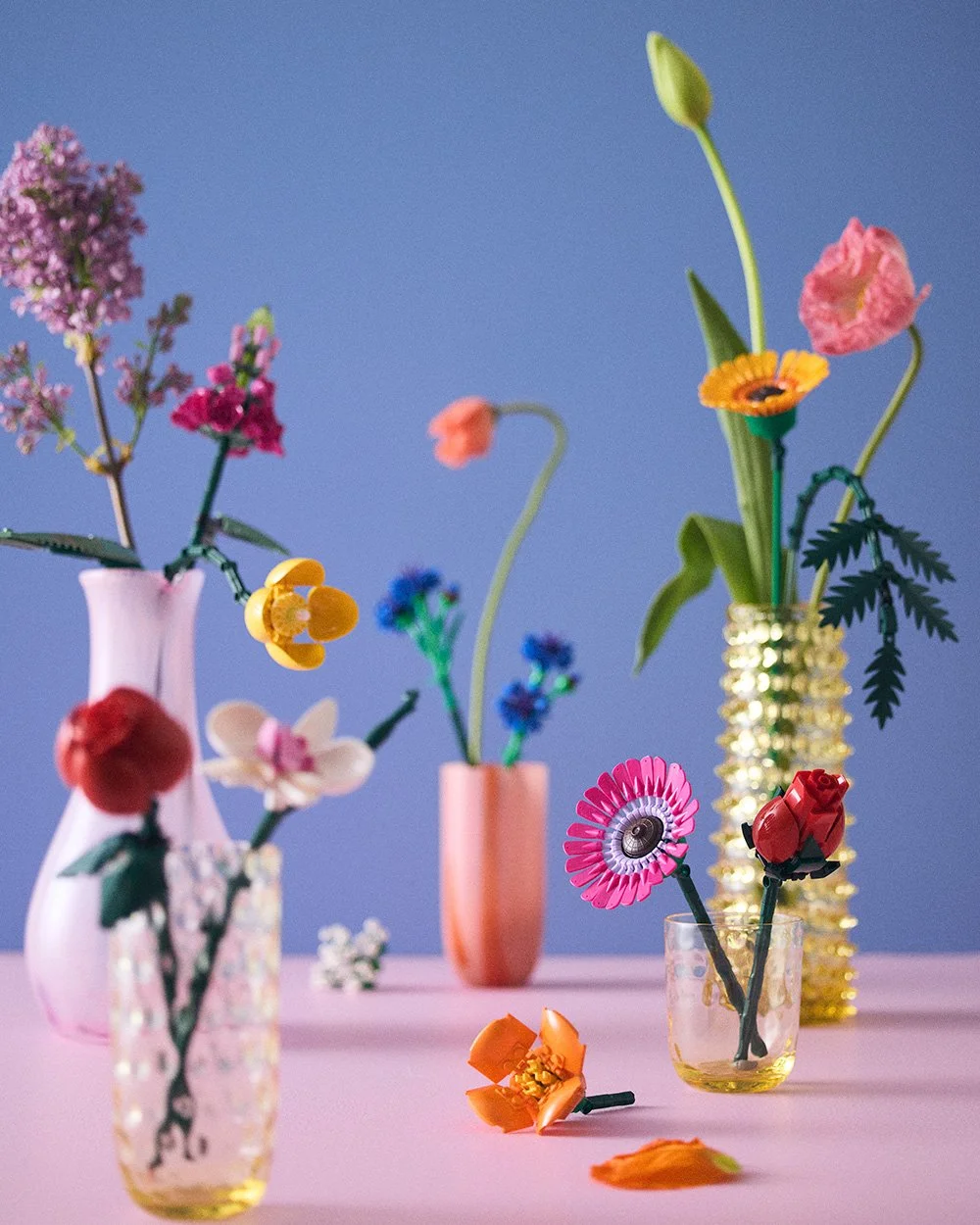Assorted colorful artificial flowers in various vases on a pink surface with a blue background.