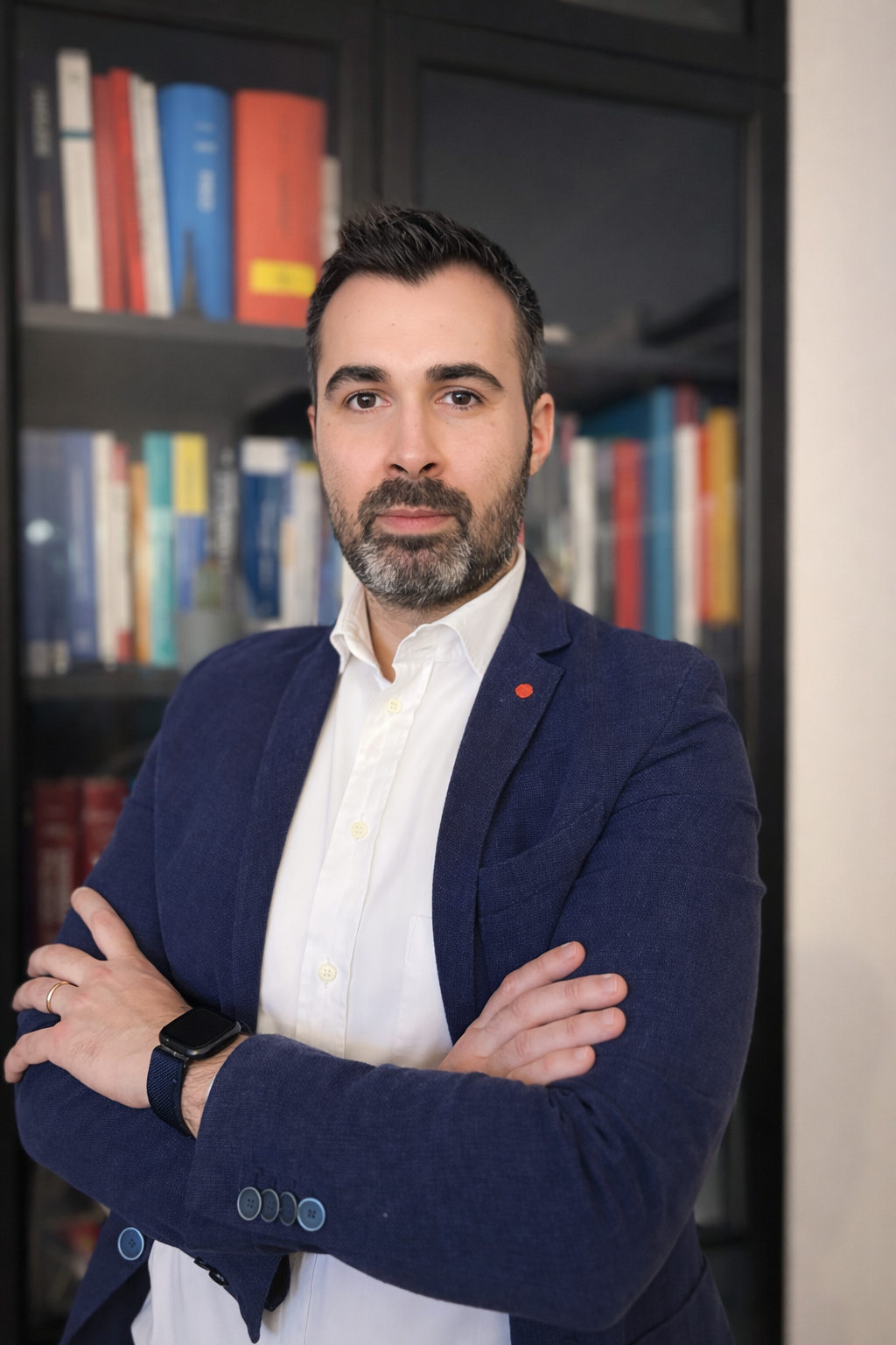 Uomo con barba e capelli scuri, indossa un blazer blu e camicia bianca, con un orologio smartwatch, in piedi di fronte a una libreria con libri colorati.