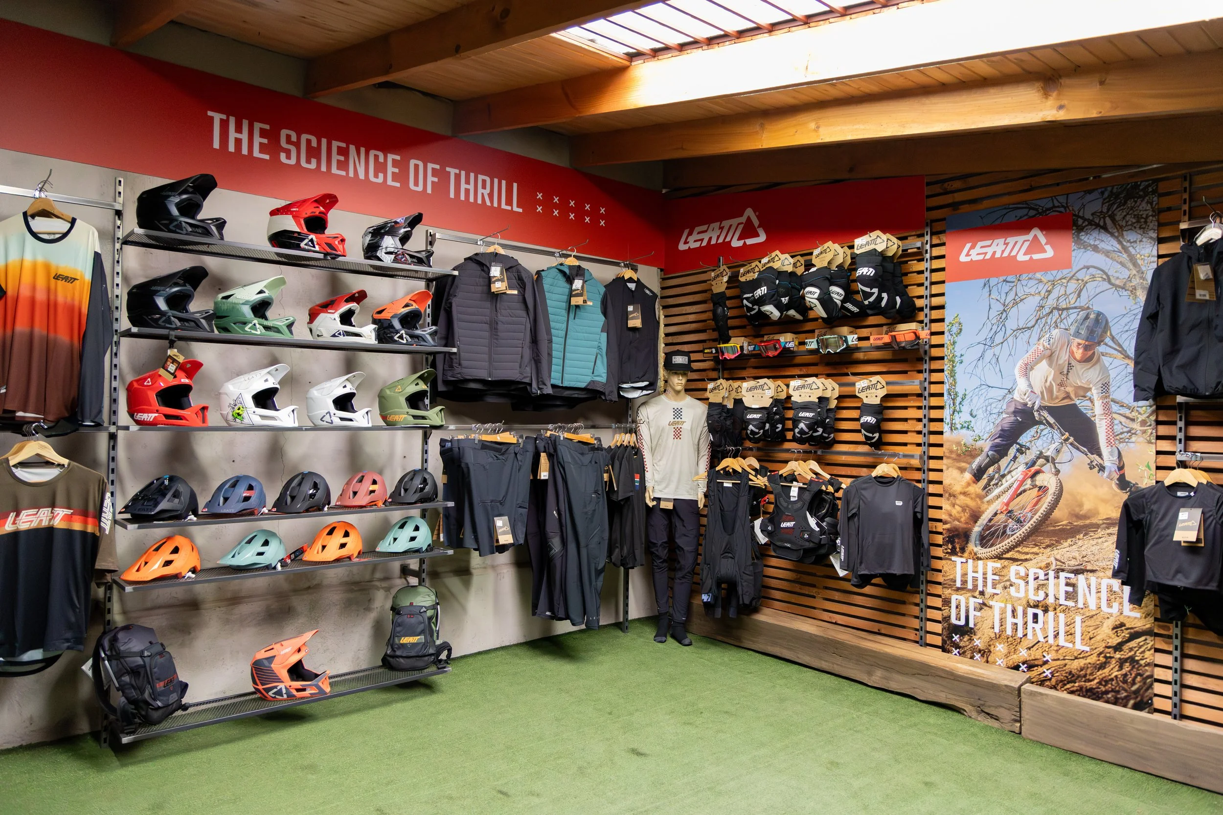 Sección de artículos de ropa y equipo para ciclismo, con cascos, chaquetas, pantalones, guantes y bolsas, en una tienda con decoración en madera y ranuras para exhibición.