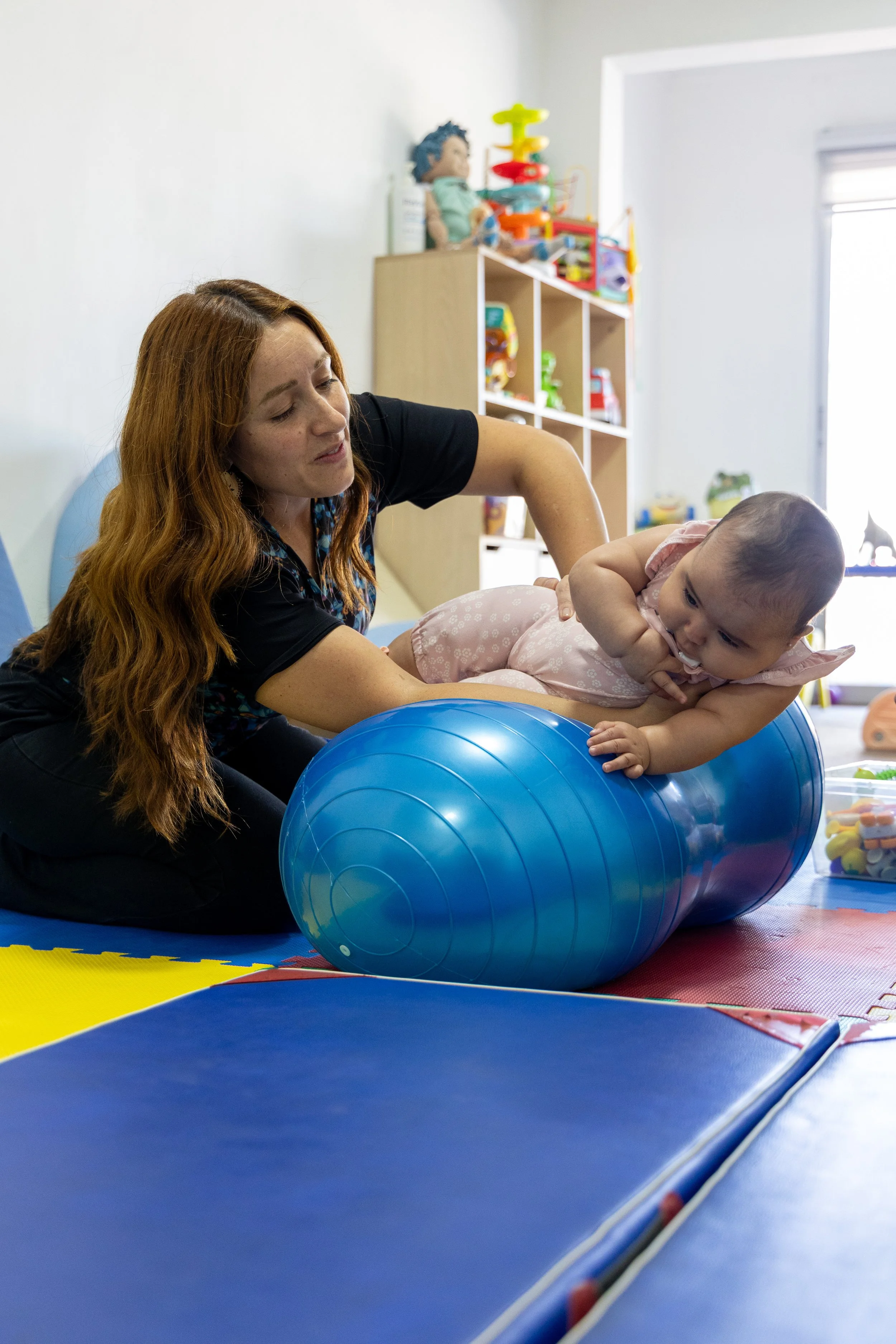 Una mujer ayuda a una niña a hacer ejercicio en una pelota azul en una sala de terapias o actividades para niños.