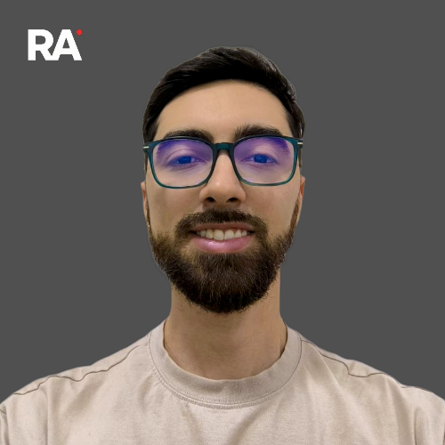 Joven con barba y gafas con marco negro y cristales azul oscuro, sonriendo, fondo gris y logotipo 'RA' en la esquina superior izquierda.