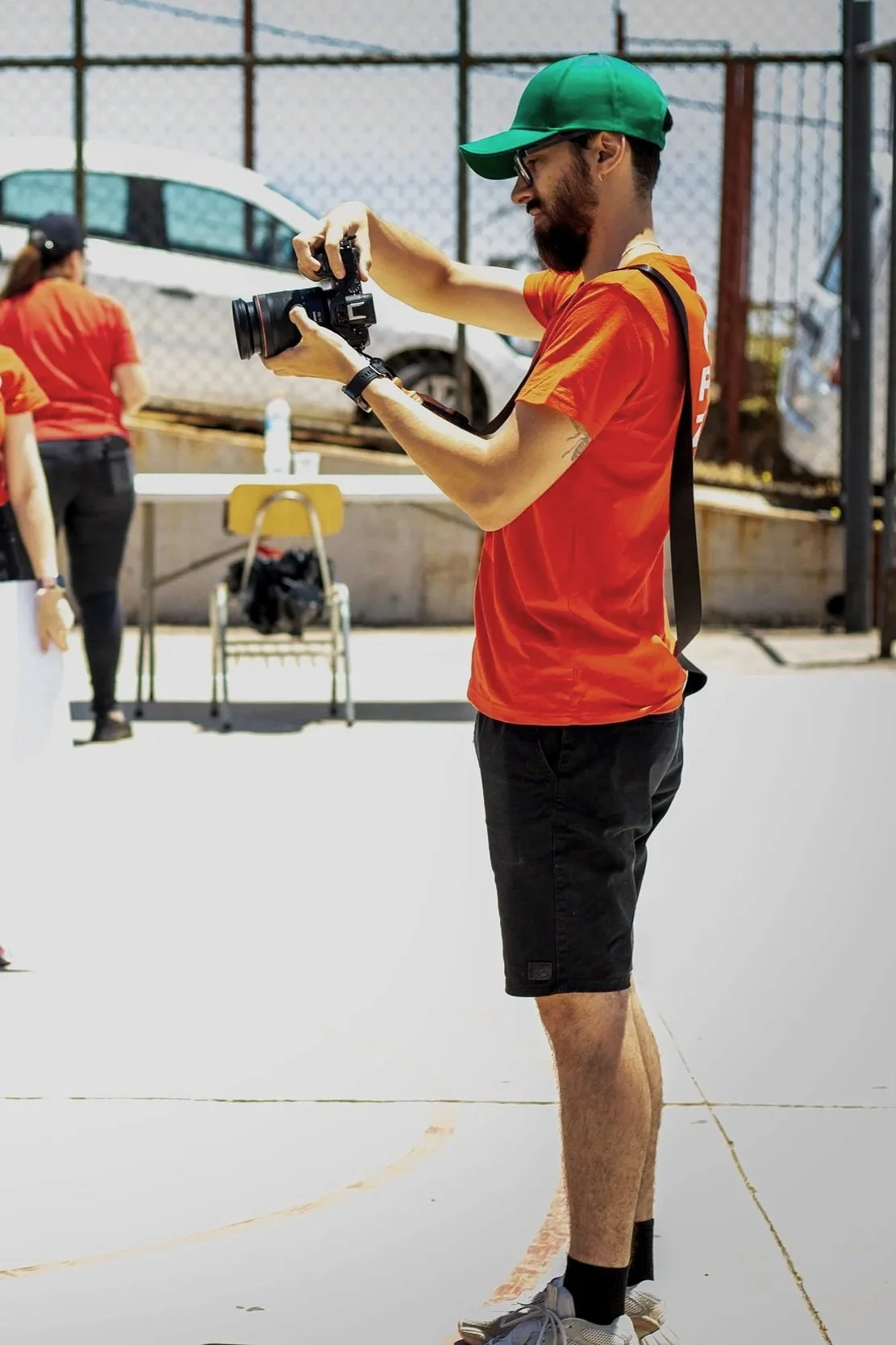 Hombre joven con camiseta roja, pantalones cortos negros, sombrero verde y gafas, tomando una foto con una cámara en un espacio al aire libre.