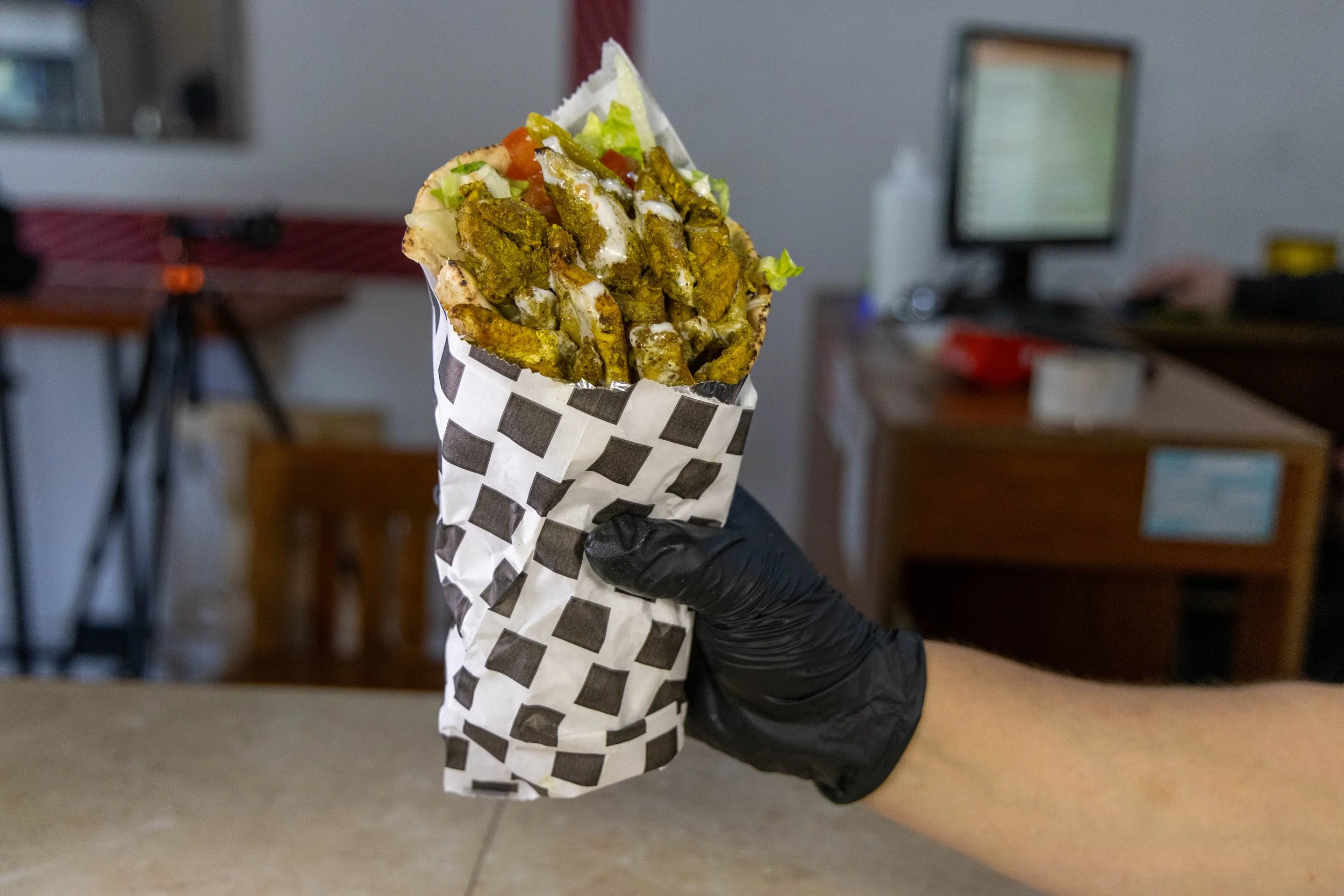 Persona con guante negro sosteniendo un panucho relleno de carne, lechuga, tomate, y salsa, envuelto en papel de cocina con cuadros blancos y negros en un establecimiento.