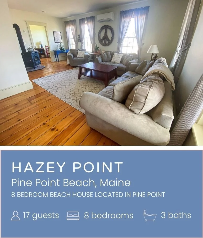 Living room with beige sofas, wooden coffee table, patterned rug, wood flooring, windows with curtains, a peace sign wall hanging, and a lamp.