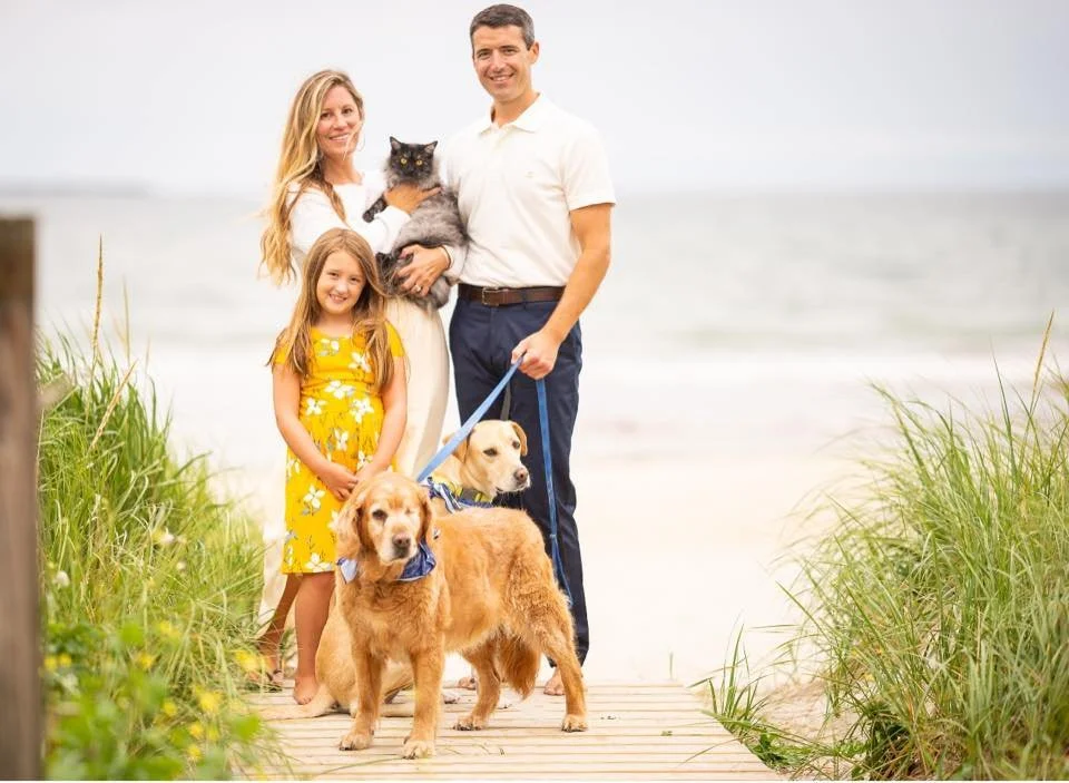 A family of four including a woman, man, young girl, and two dogs, with a cat, standing on a wooden walkway leading to the beach, during daytime