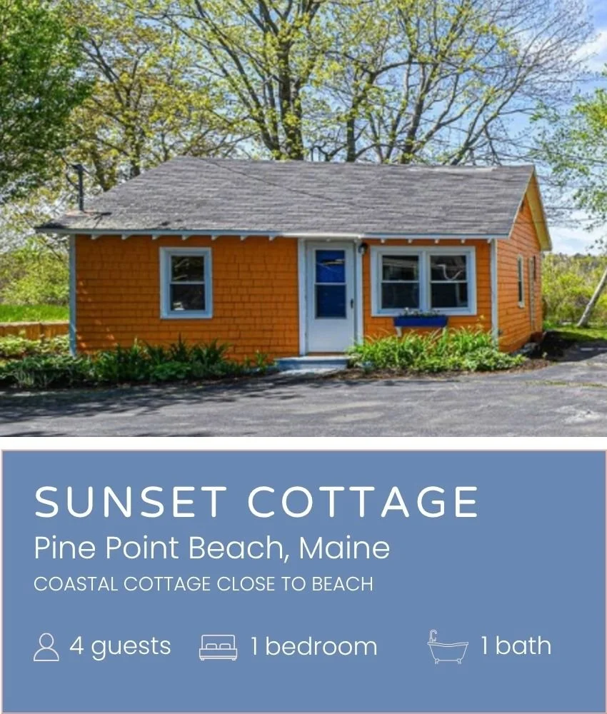 A small, orange wooden cottage with a gray shingled roof, white trim around the windows and door, and surrounded by green plants and trees.
