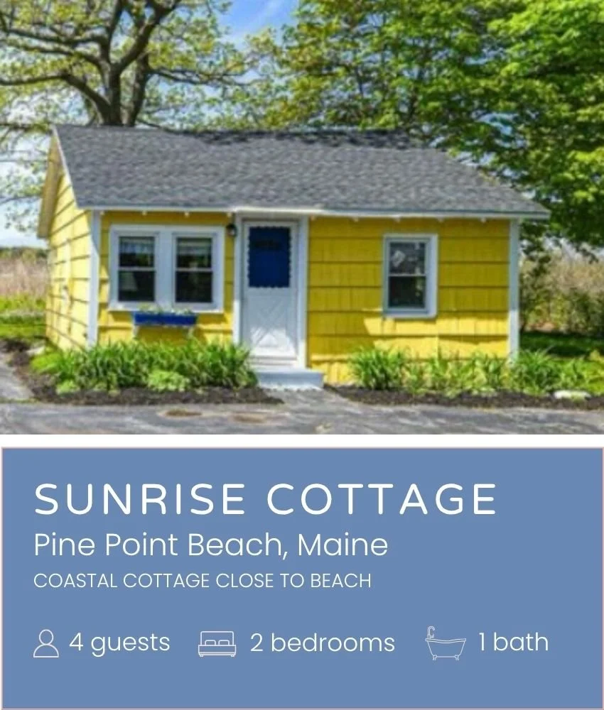 Bright yellow cottage with white window trim and gray shingled roof, surrounded by green plants and trees in a sunny outdoor setting.
