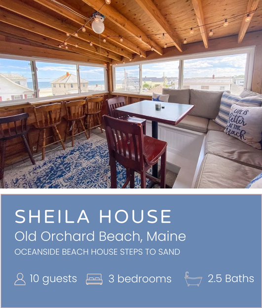 Cozy sunroom with wooden ceiling, string lights, and large windows showing beach houses and the ocean in Old Orchard Beach, Maine. Contains a dining table, chairs, and a built-in seating area with cushions.