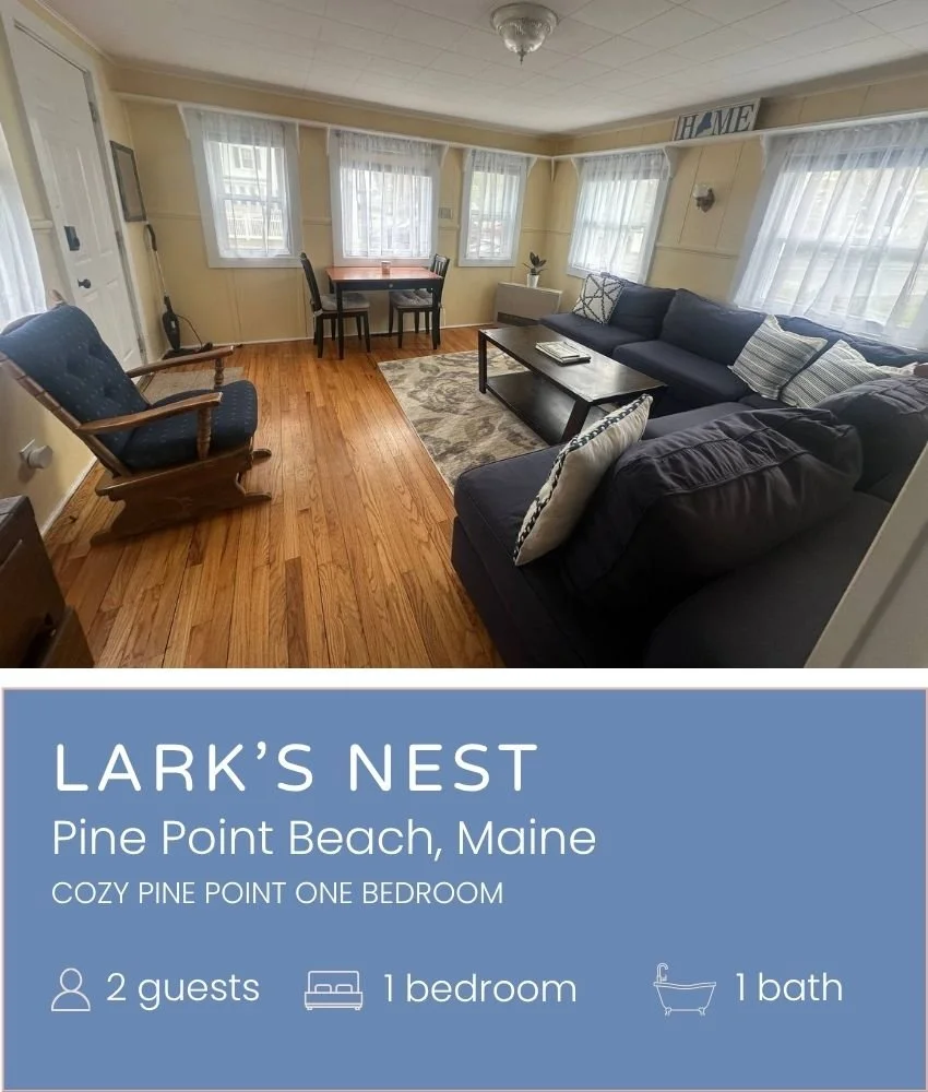 Cozy living room with four windows, a black sectional sofa with pillows, a wooden coffee table, a wooden rocking chair, a small dining table with chairs, and a rug on hardwood floors in a beach house.