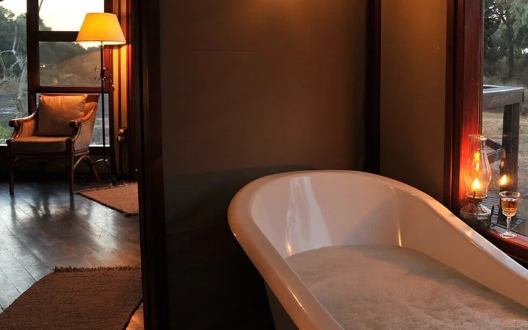 A cozy room with a bathtub filled with water, a lit lamp, and a chair next to a window showing an outdoor scene with trees and a wooden deck.