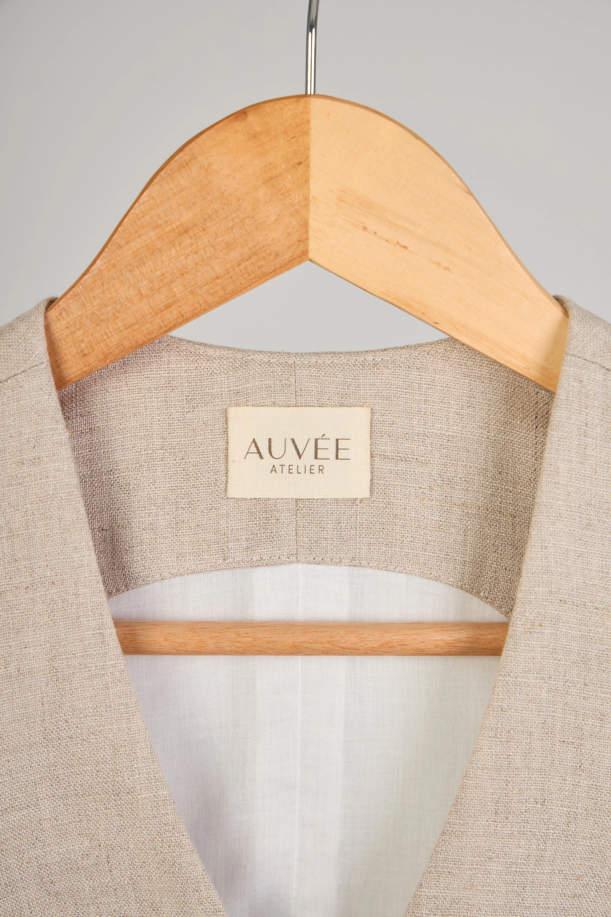 Close-up of a beige linen jacket hanging on a wooden hanger with a label reading "AUVÉE ATELIER" inside.
