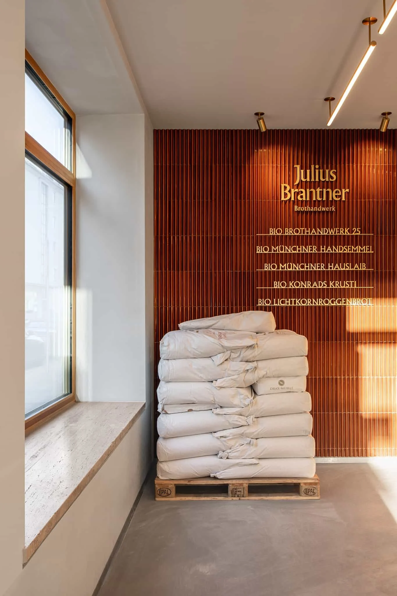 Ein Stapel mit weißen Säcken auf einer Palette in einem Raum mit warmer Holzvertäfelung an der Wand, die den Namen "Julius Brantner" und eine Liste von Bio-Backwaren zeigt.