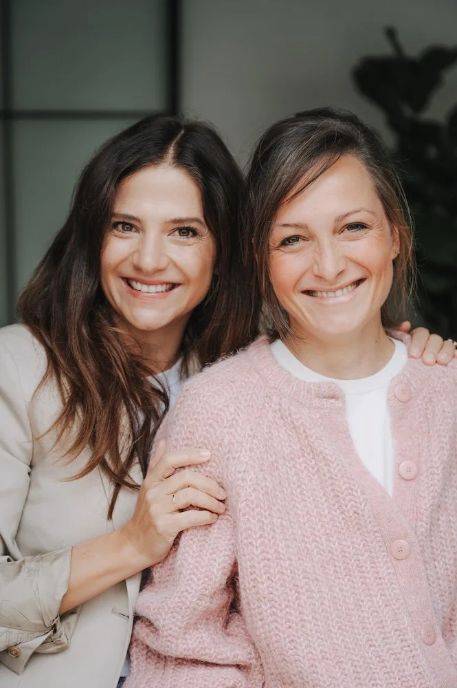 Zwei lachende Frauen, eine in einem beigen Blazer und die andere in einer rosa Strickjacke, stehen eng beieinander und posieren für die Kamera.