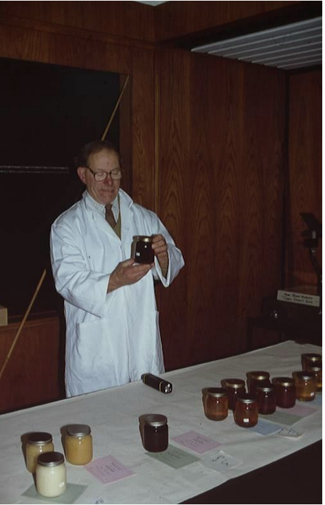 A man in a white lab coat taking a photo of a display with various honey jars on a table, in a wooden-paneled room