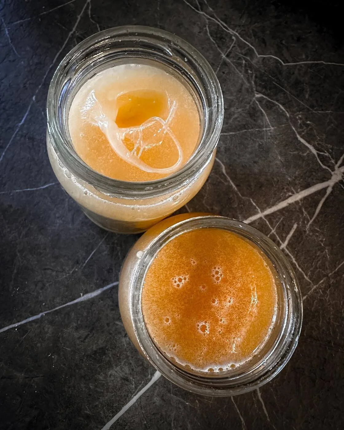 Two mason jars filled with orange-colored beverages placed on a dark marble surface, one with ice cubes inside.