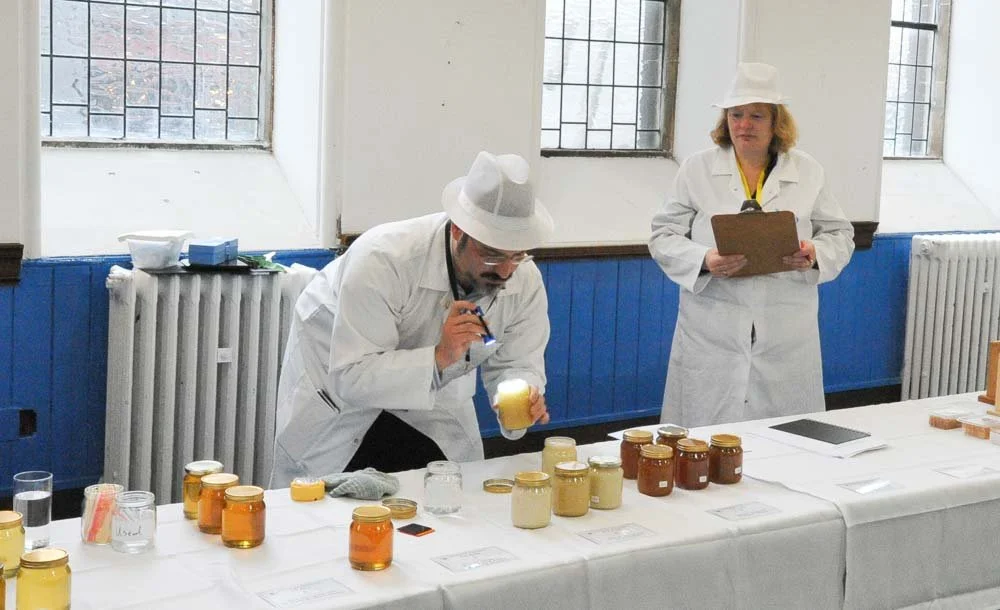 Two people in white lab coats and hats examining jars of honey at a table, with one person taking notes on a clipboard.