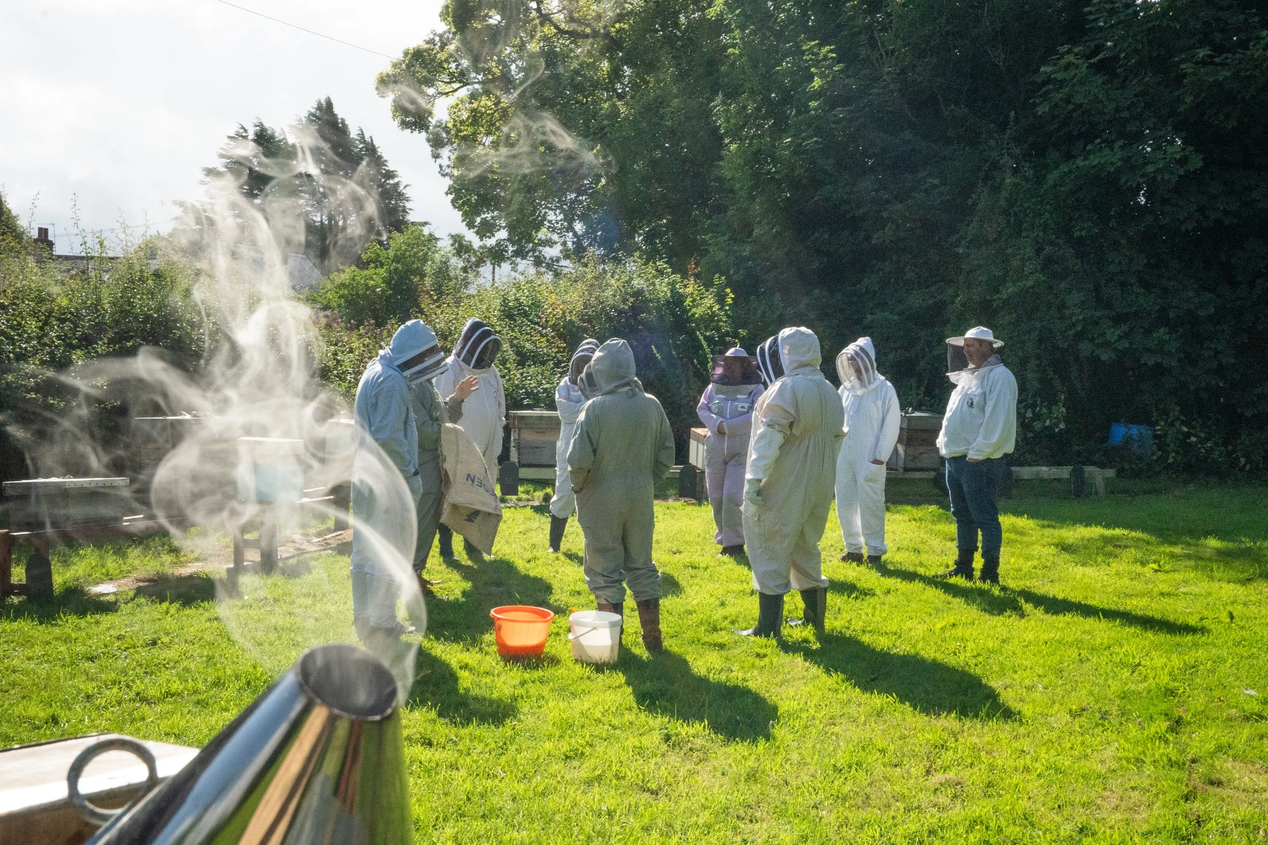 A group of beekeepers in protective suits and veils are gathered outdoors on a grassy field, with trees in the background, during daytime. A steaming bee smoker is visible in the foreground.