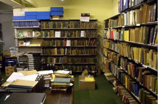 A room with many bookshelves filled with books and documents, some stacked on tables and the floor. The Moir Library, Edinburgh.