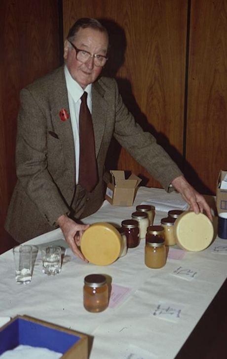 An elderly man in a brown suit and glasses standing at a table with jars of honey and other items. He is holding a jar, smiling at the camera.