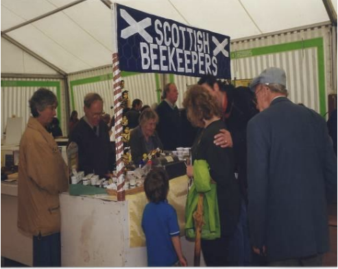 People gathered at a Scottish Beekeepers booth inside a tent, with a sign displaying "Scottish Beekeepers" and various items on the booth counter. The Royal Highland Show.