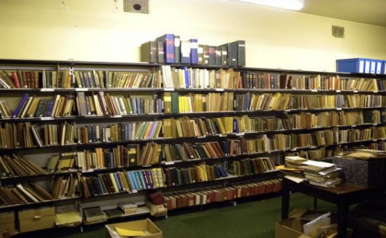 A bookshelf filled with books and binders in a room with green carpet and beige walls. The Moir Library, Edinburgh.