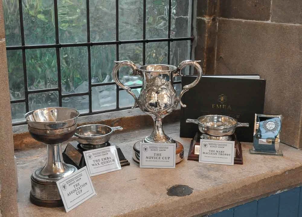 A display of silver trophies and awards on a windowsill, including the Novice Cup, the EMBA Wax Quiche, and the Mary Maclean Trophy, with a book.