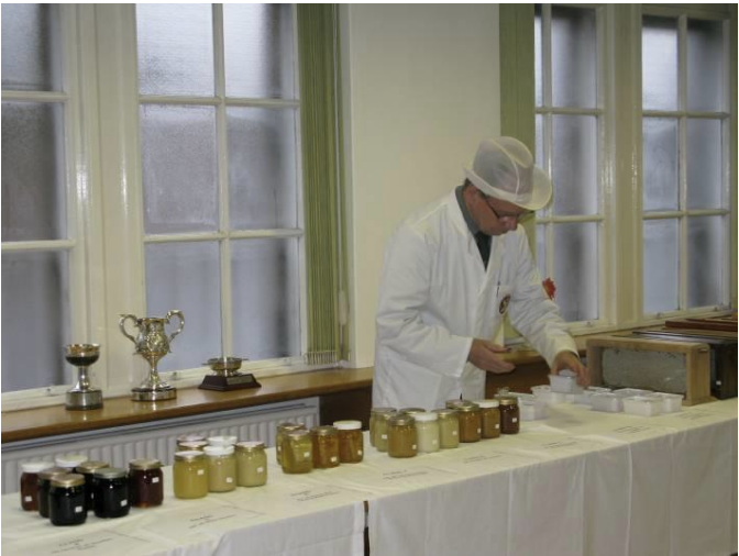 Person in white coat and cap examining jars of honey on a table in a room with large windows.