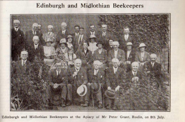 Group photo of Edinburgh and Midlothian beekeepers at the apiary, including men and women in old-fashioned clothing, some holding hats, taken outside with greenery in the background.