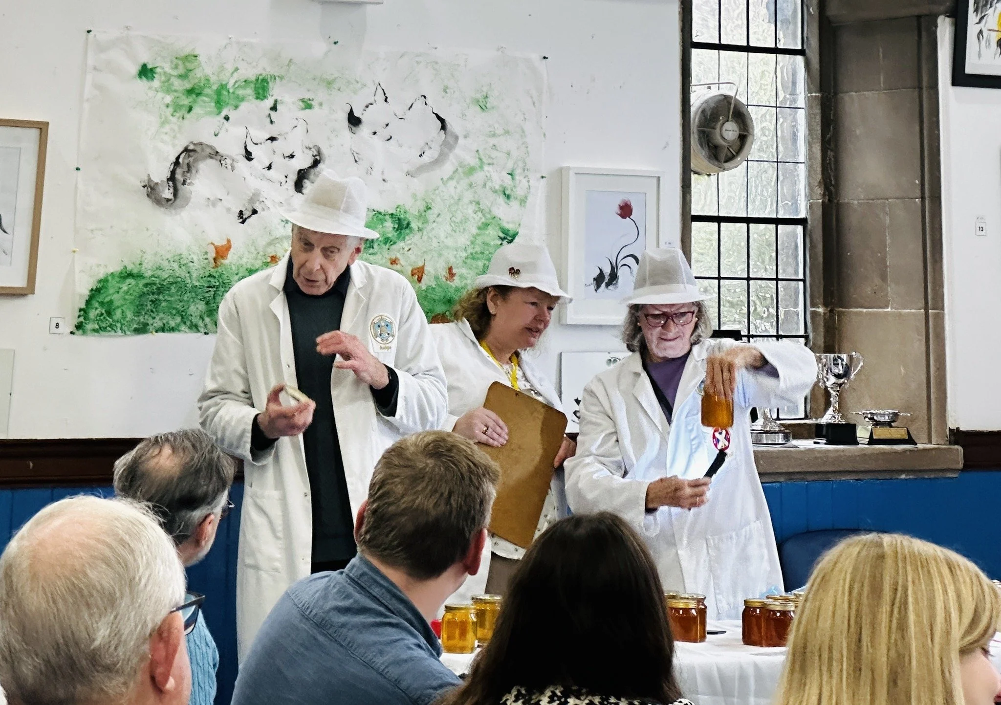 Three people dressed as scientists or chemists giving a presentation or demonstration to an audience, with jars of honey on the table in front of them. They are wearing white lab coats and hats, and are inside a room with artwork and large windows.