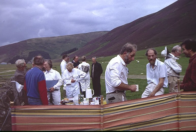 Group of beekeepers having an outdoor gathering or picnic in a grassy field with heather hills in the background. Some are standing and chatting, with tables and supplies visible in the foreground.