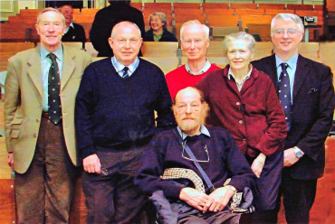 Group of six people, five standing and one sitting, posing in a room with wooden wall and conference seating.