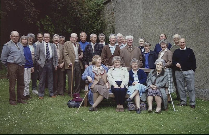 A group of people outdoors on grass, posing for a photo with some sitting and others standing under trees and beside a wall.