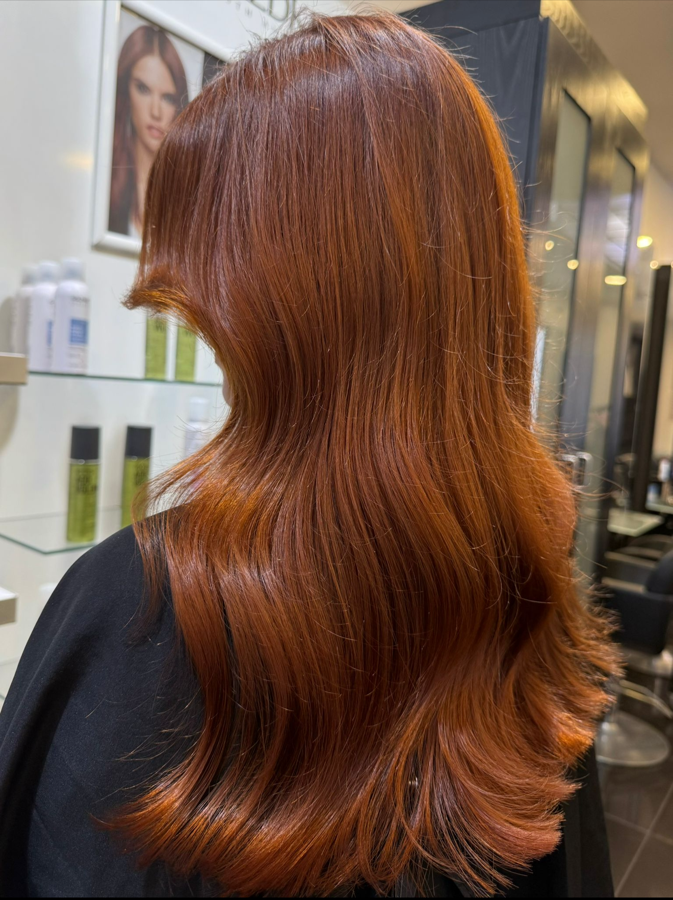 Back view of a woman with long, wavy, vibrant red hair styled with layers, in a salon.