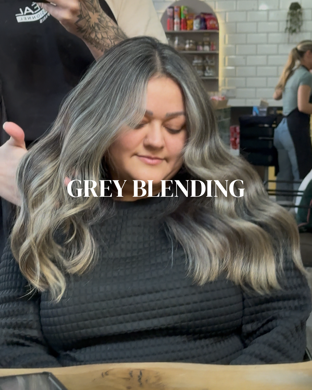A woman with long, wavy, grey-blended hair sitting in a salon with her eyes closed. The text 'GREY BLENDING' overlays the image.