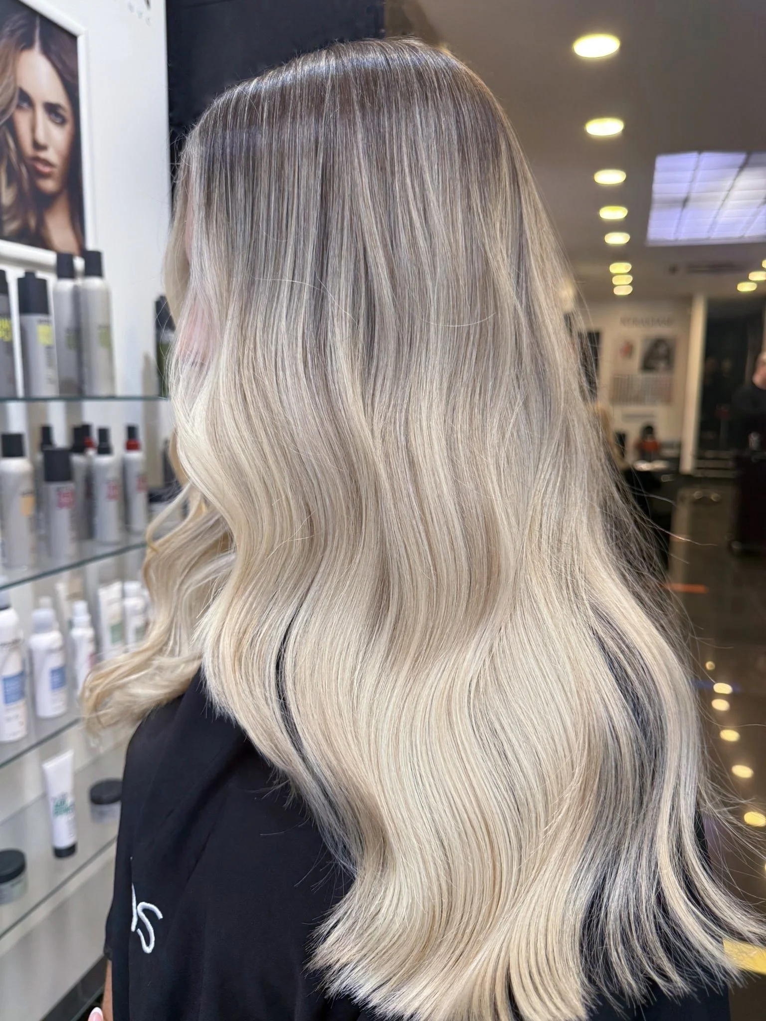 A woman with long, wavy, blonde hair in a salon setting, with shelves of hair products in the background.