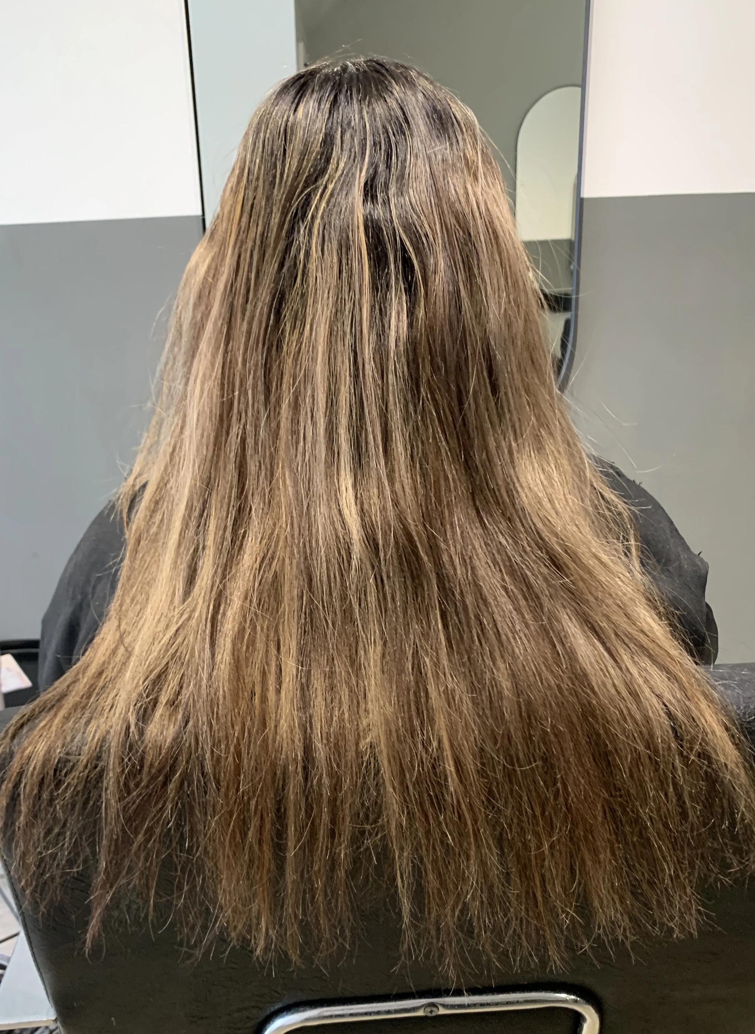 Back of a person's head with long, straight, brown hair, sitting in a salon chair, with a mirror and gray walls in the background.