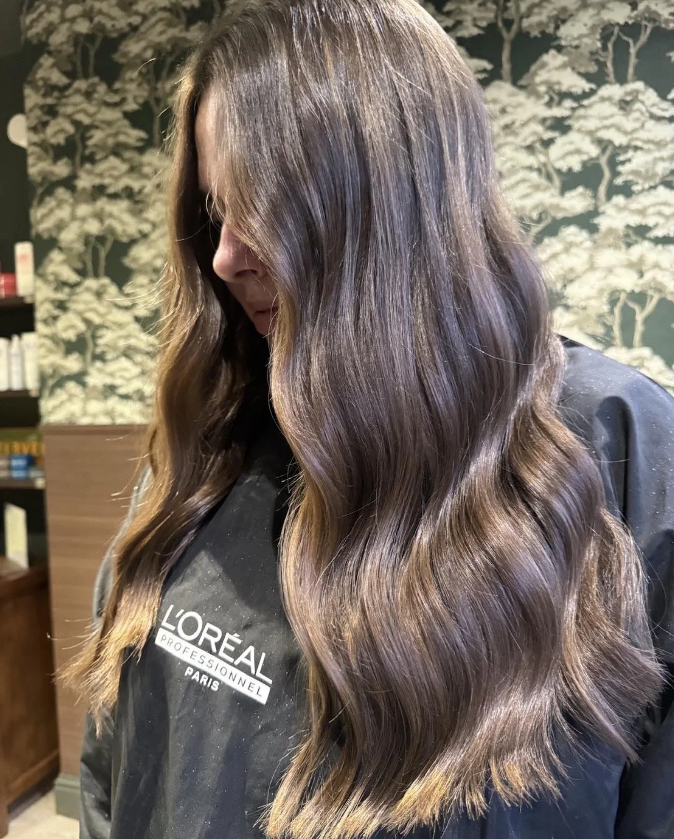 A woman with long, wavy, brown hair looking down, wearing a black L'Oreal professional jacket, inside a salon with wallpaper featuring white clouds and trees.