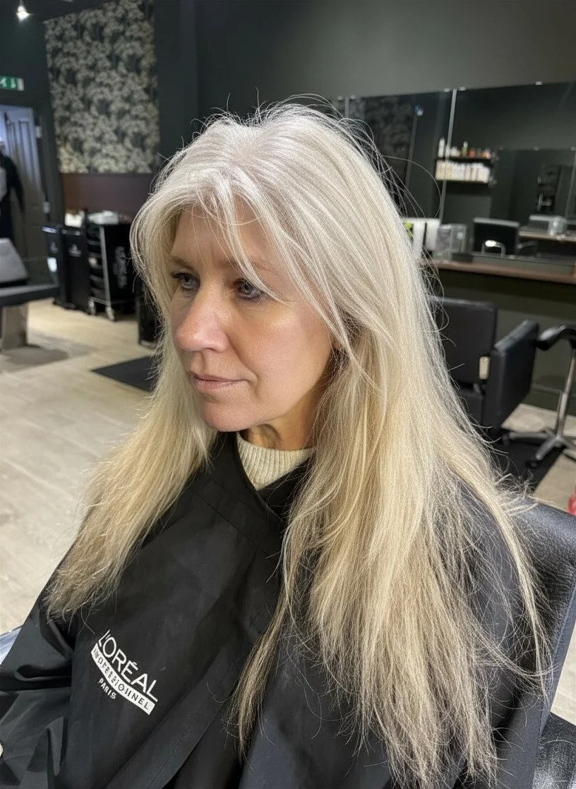 A woman with long, blonde hair and a neutral expression sitting in a salon chair, wearing a black salon cape labeled L'Oreal Professionnel, inside a modern hair salon.