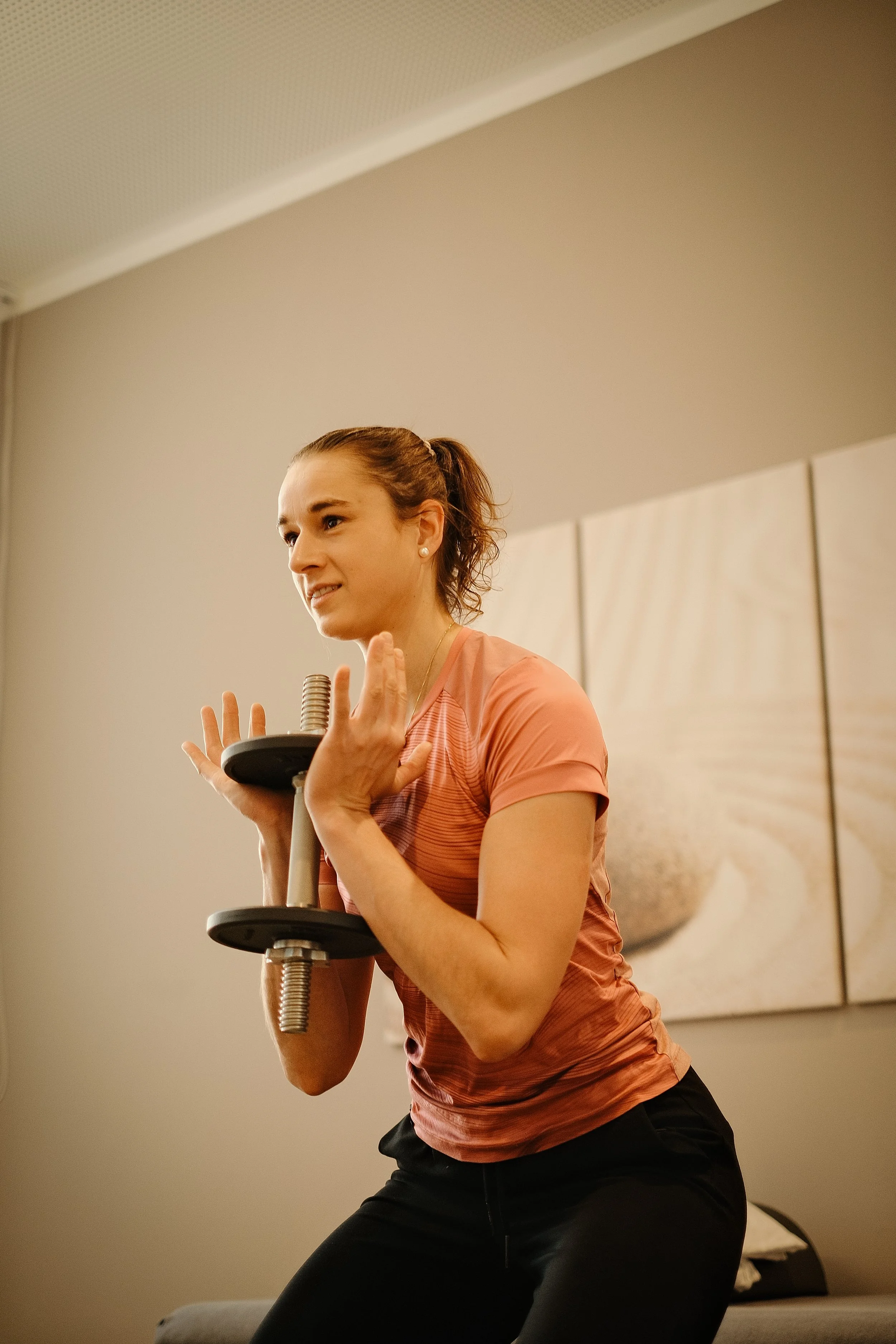 A woman is exercising indoors, holding a dumbbell in one hand, wearing a pink workout shirt and black pants, with a neutral-colored wall and a bed in the background.